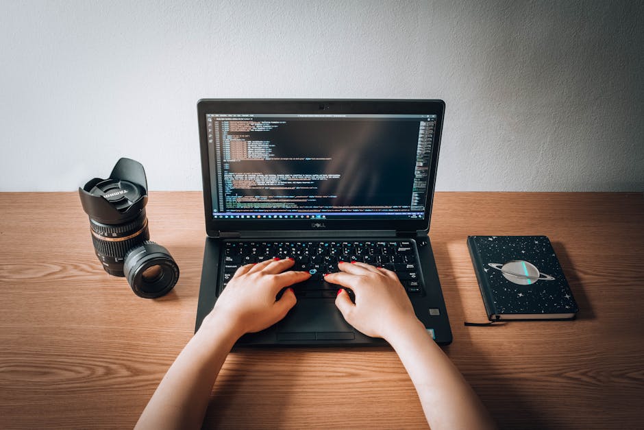 A person with short dark hair seen from behind working at a desk in an office environment, focusing on a computer monitor displaying lines of computer code with syntax highlighting in various colors. 