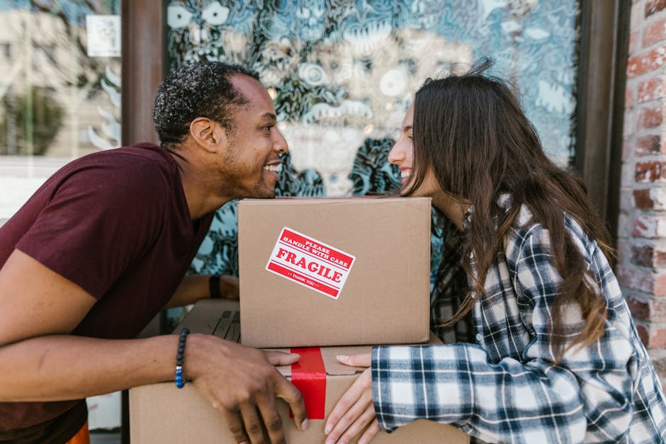 A man and a woman smile at each other while holding stacked cardboard boxes, with the top box labeled 'Fragile' and 'Handle with Care'. They are positioned outdoors in front of a large window with dec